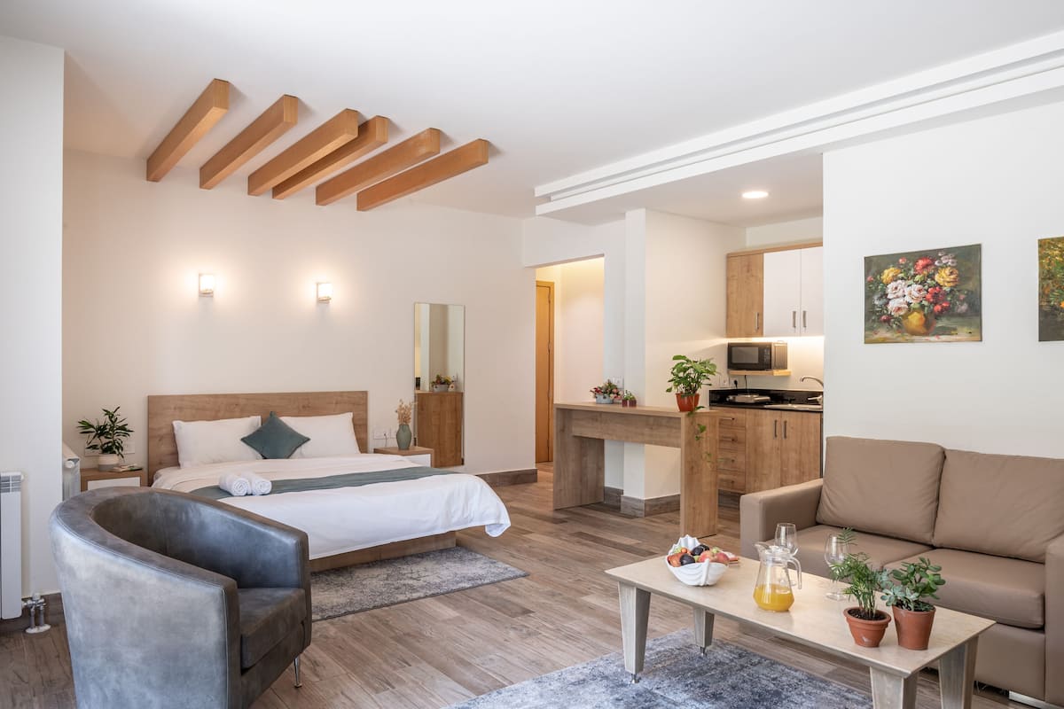A modern hotel room with a wooden ceiling, a bed with white linens, a grey armchair, a wooden coffee table with a fruit bowl, and a kitchen area in the background.