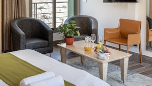 Ein Hotelzimmer mit Bett, zwei Sessel, einem Tisch mit Pflanze und Getränken, einem Fernseher und Blick ins Freie.
