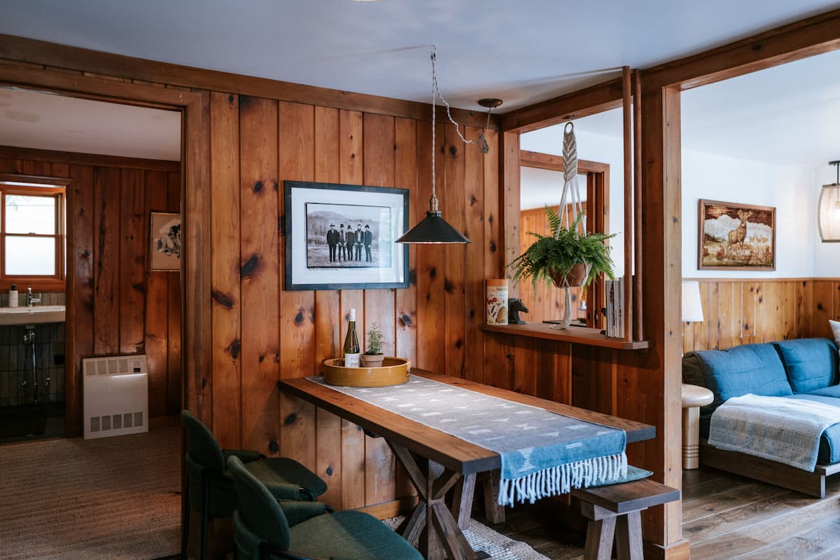 A rustic interior with wooden walls, a dining table with a bench, a sofa, and a hanging light fixture.