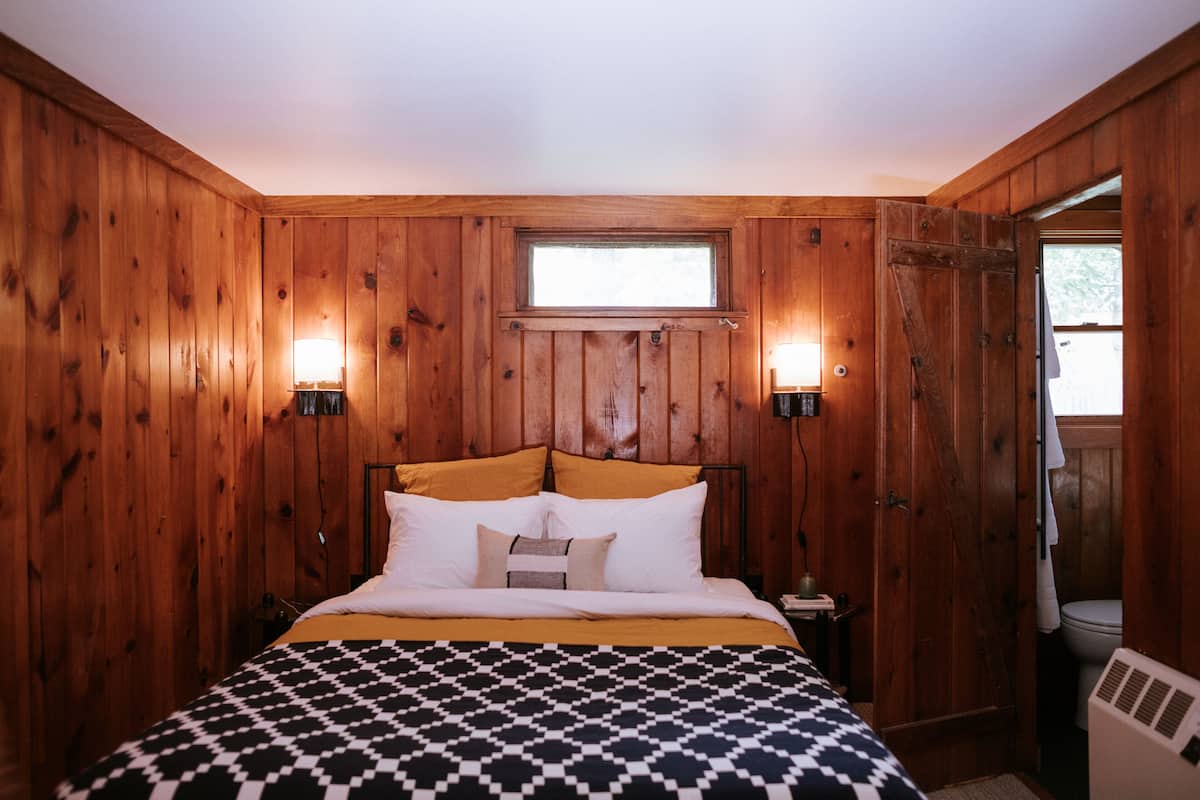 A wooden-paneled bedroom with a bed, two bedside lamps, a window, and a door leading to a bathroom.