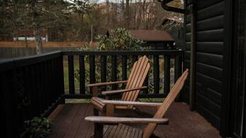 Een houten terras met twee Adirondack-stoelen, een zwarte reling en een uitzicht op bomen en een gebouw op de achtergrond.