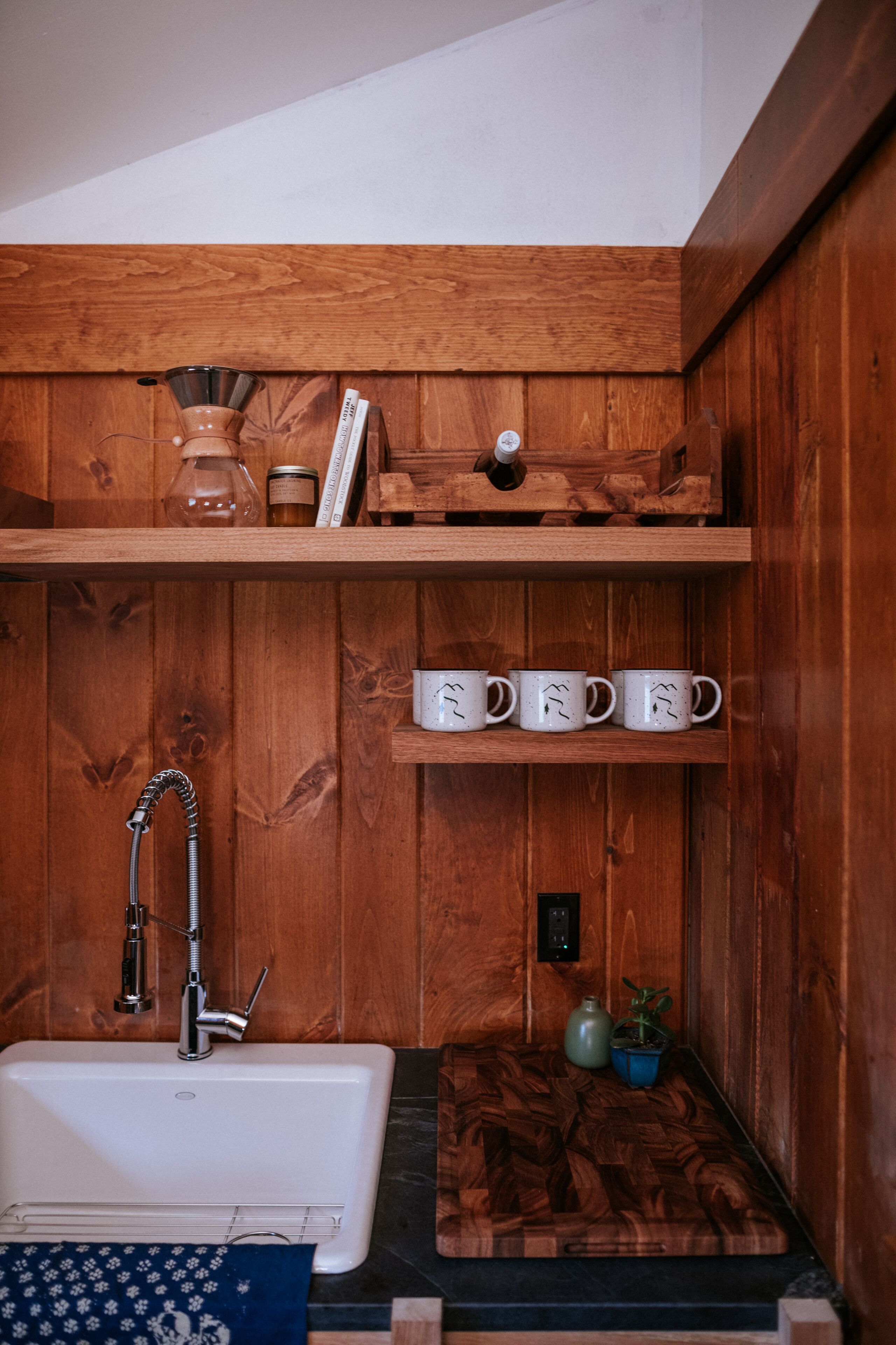 Un baño de madera con lavamanos blanco, ducha y estante con tazas y una planta.