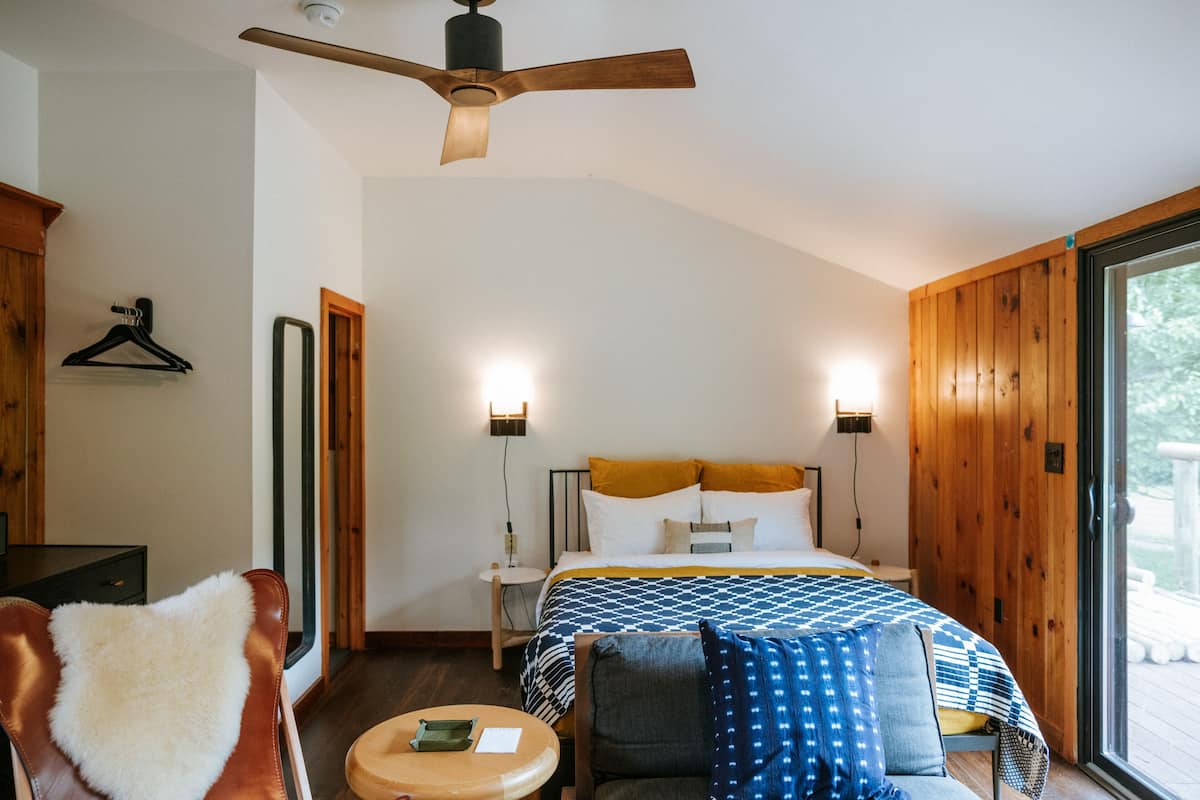 A bedroom with a bed, a chair, a table, a ceiling fan, and a window.