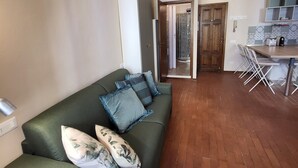 A living area with a green sofa, a wooden ceiling beam, a kitchen with a tiled backsplash, and a door leading to a bathroom. - 40 m2 studio in the center of Brescia, 90 m from parking, no ZCL (Brescia)