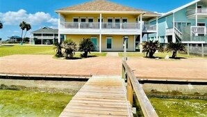 A two-story building with a balcony, a wooden dock leading to a water body, and palm trees. - Wake Up to a Beautiful View! 3bdrm, 2 bath. Private Fishing Pier. (Port Mansfield)