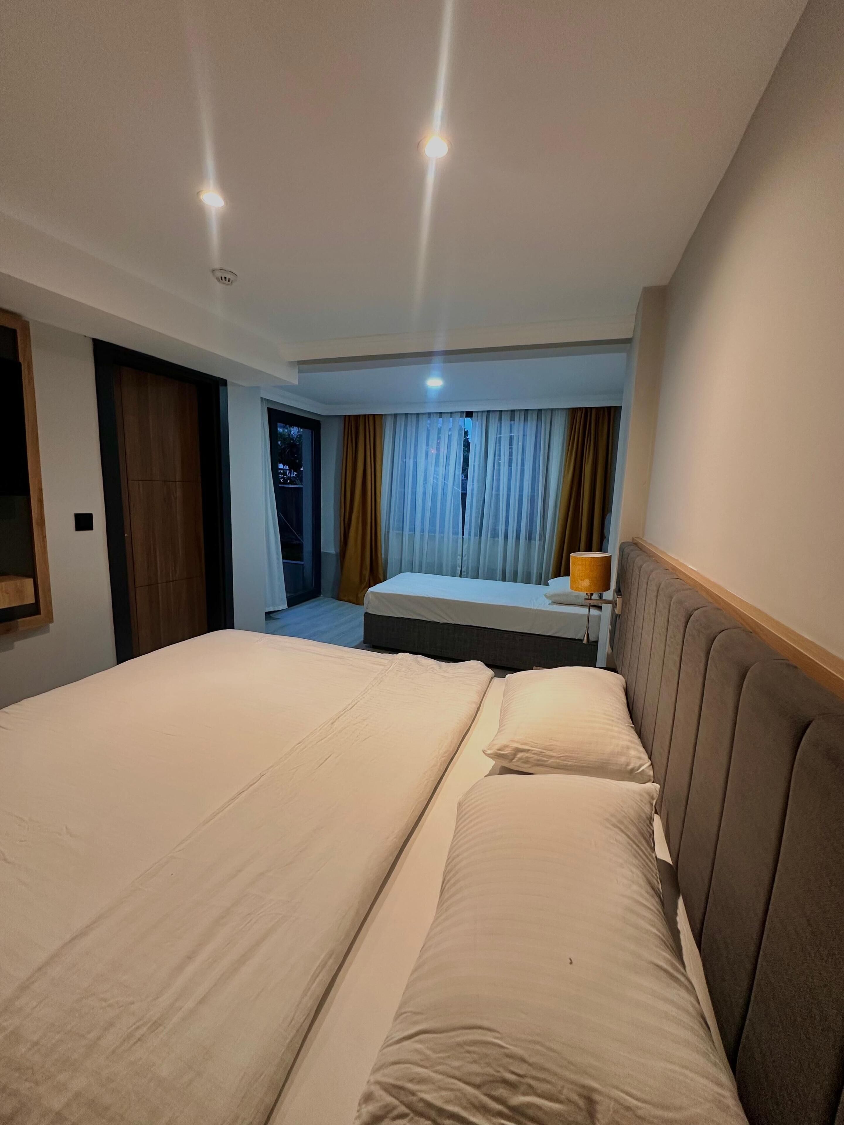 Une chambre d’hôtel avec un grand lit, un plus petit lit, une table de chevet avec une lampe, une armoire et une fenêtre avec des rideaux.
