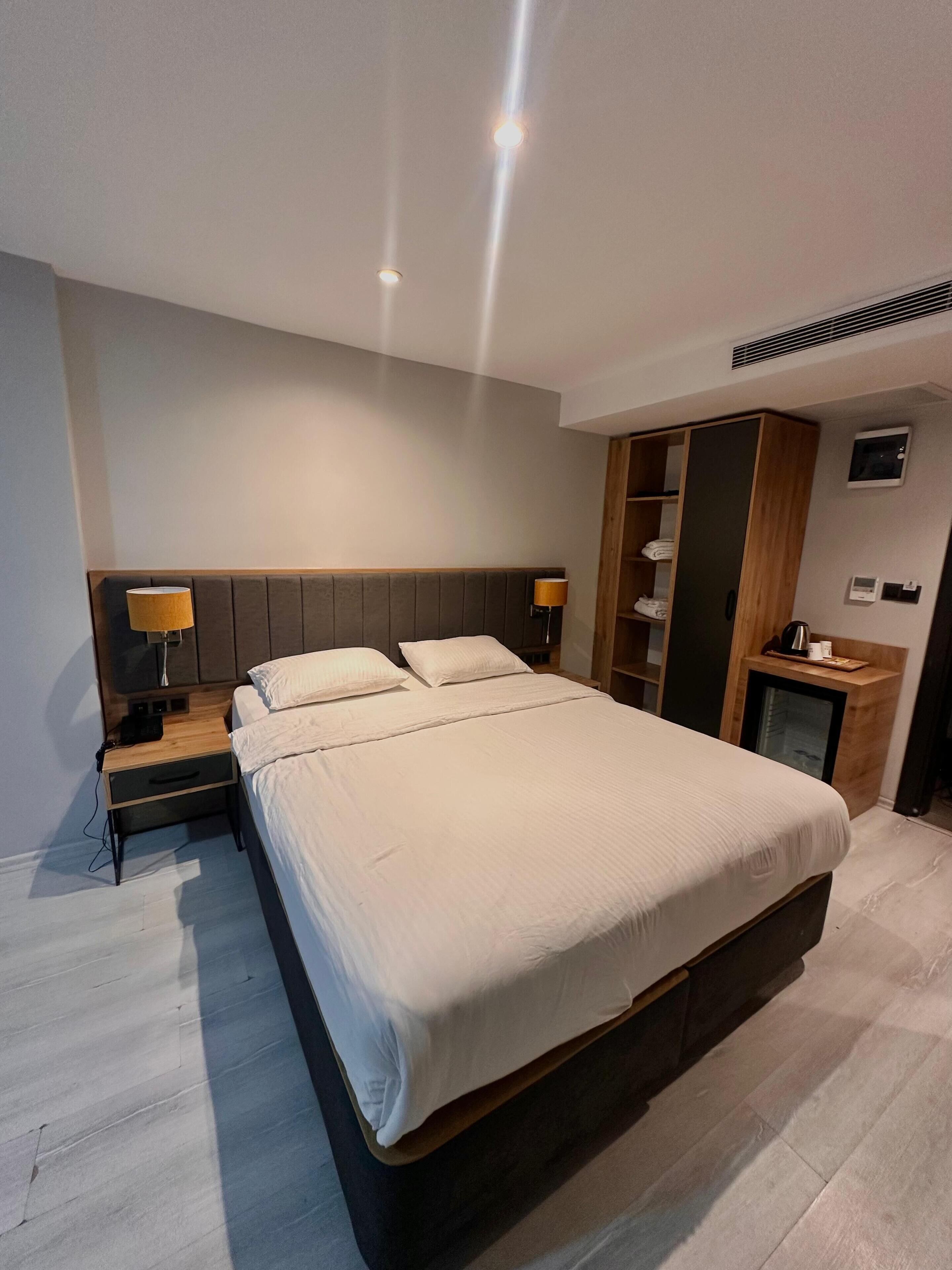 A modern hotel room with a large bed, bedside lamps, a wardrobe, and a small desk with a kettle.