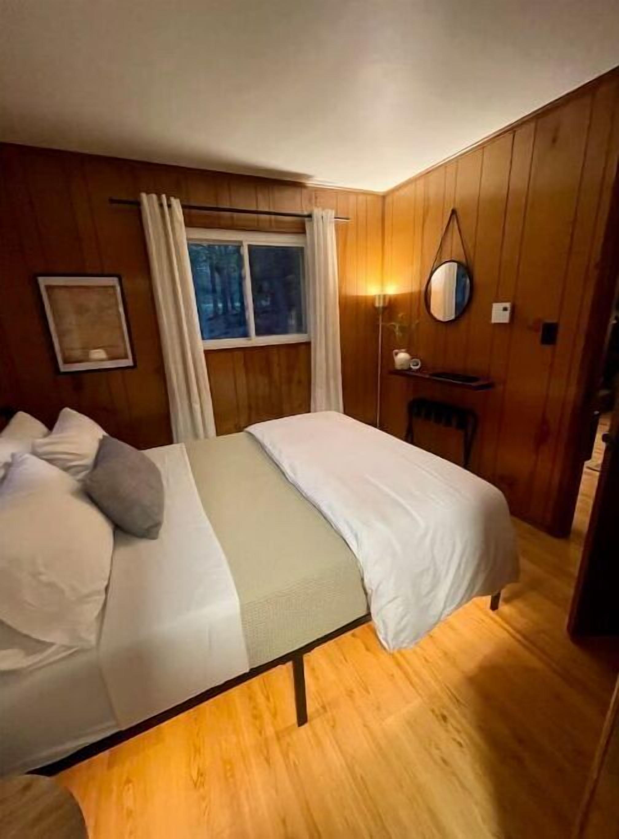 A bedroom with a bed, a bedside table, a mirror, and a window with curtains.