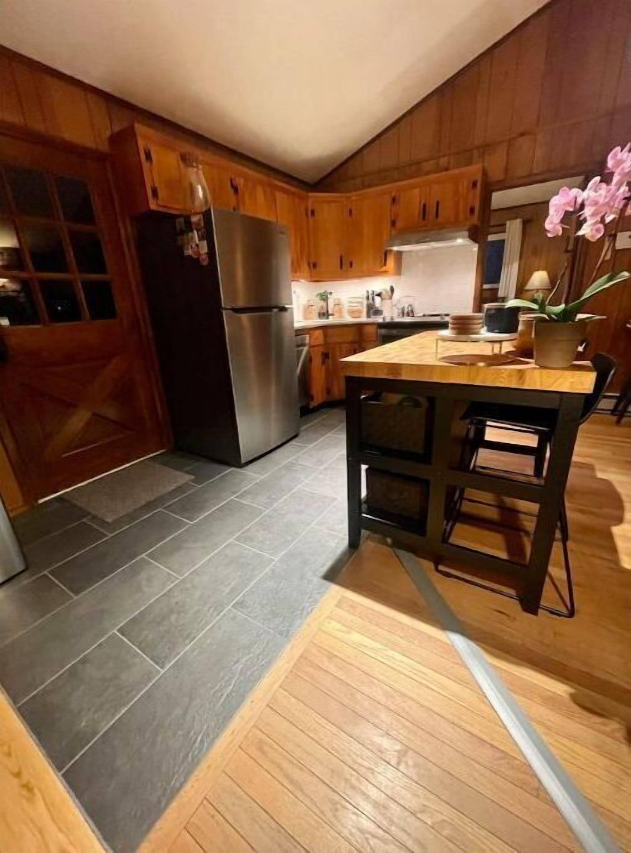 A kitchen with wooden cabinets, a stainless steel refrigerator, a wooden island with stools, and a potted orchid.