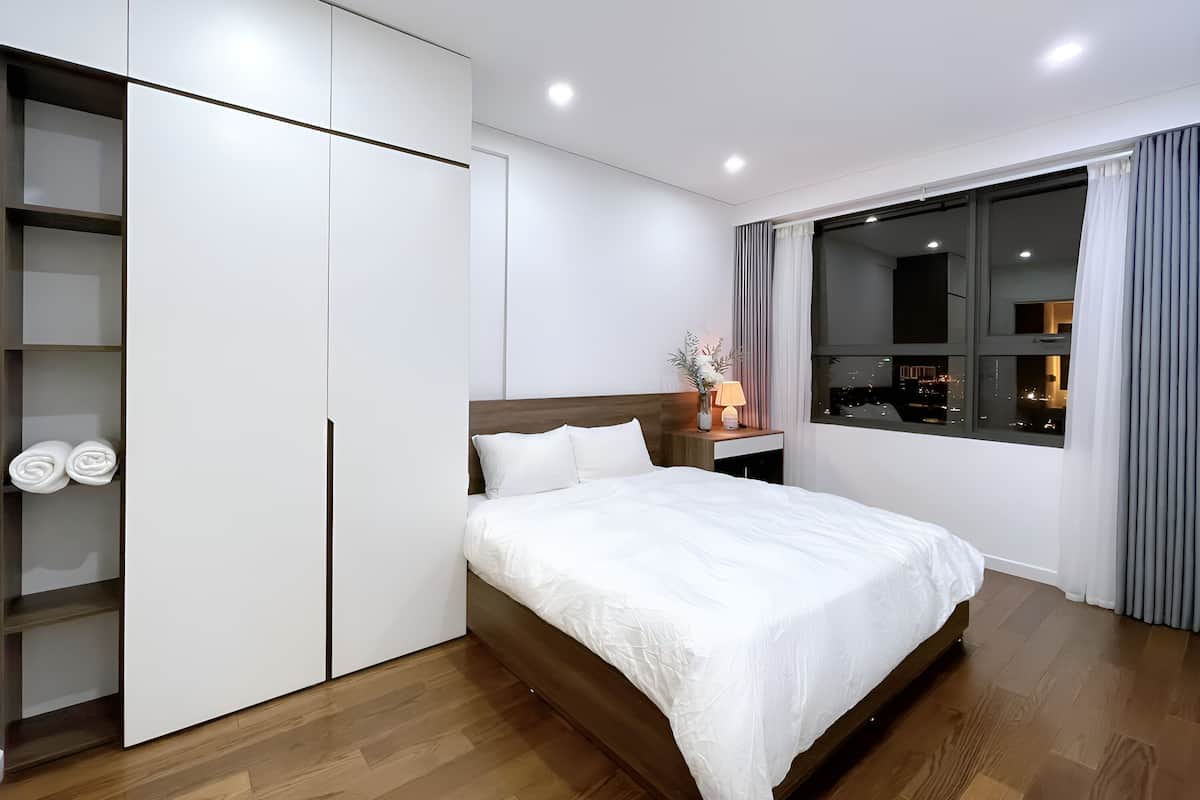 A modern hotel room with a large bed, a wardrobe, a nightstand with a lamp, and a window with a city view.