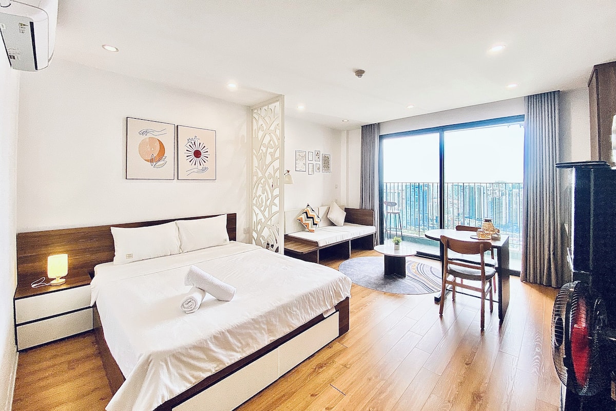 A hotel room with a bed, bedside table, and a small desk with a chair. There is a large window with a view of the city.