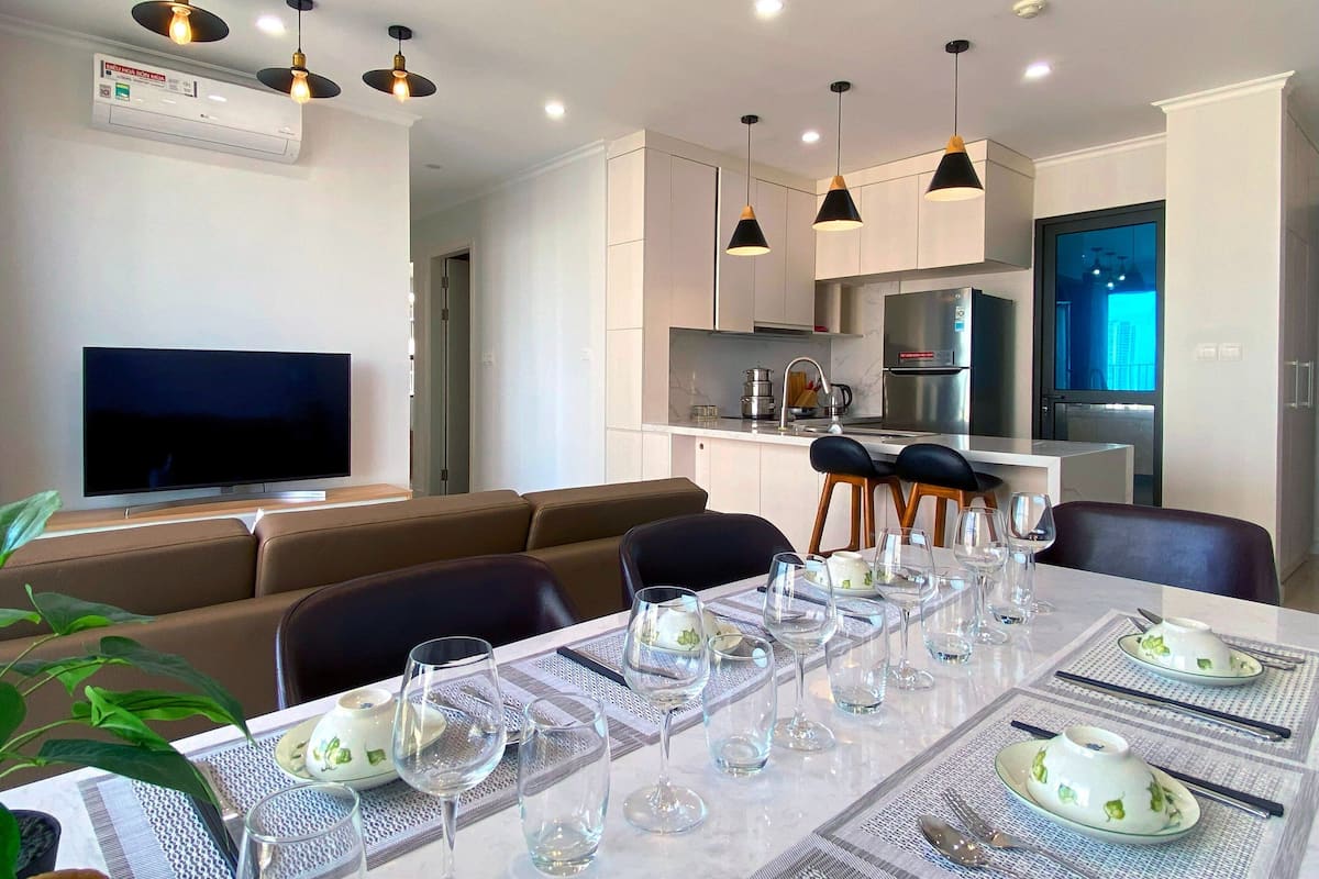 A modern living room with a flat-screen TV, a dining table set for six with glasses and plates, and a kitchen area with a refrigerator and stove.