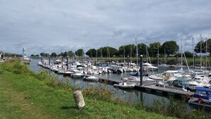 Un port de plaisance où de nombreux bateaux sont amarrés le long du rivage, une zone herbeuse avec une structure en pierre et un ciel nuageux.