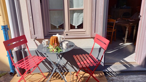 Une petite table d’extérieur avec deux chaises rouges, une théière blanche et deux tasses blanches, le tout posé sur une terrasse en bois.