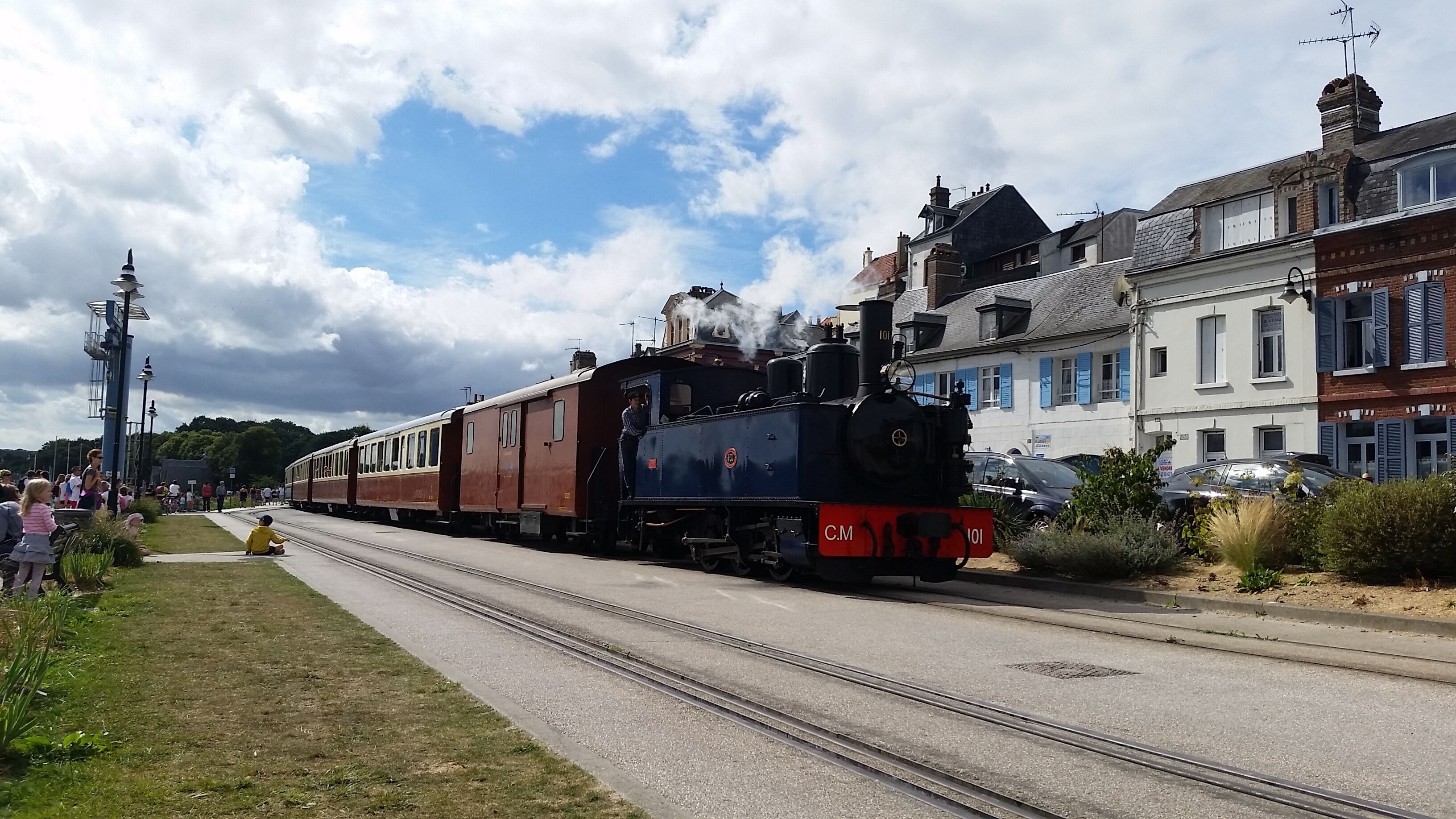 Een oude stoomtrein op rails, met een menigte mensen en gebouwen op de achtergrond.