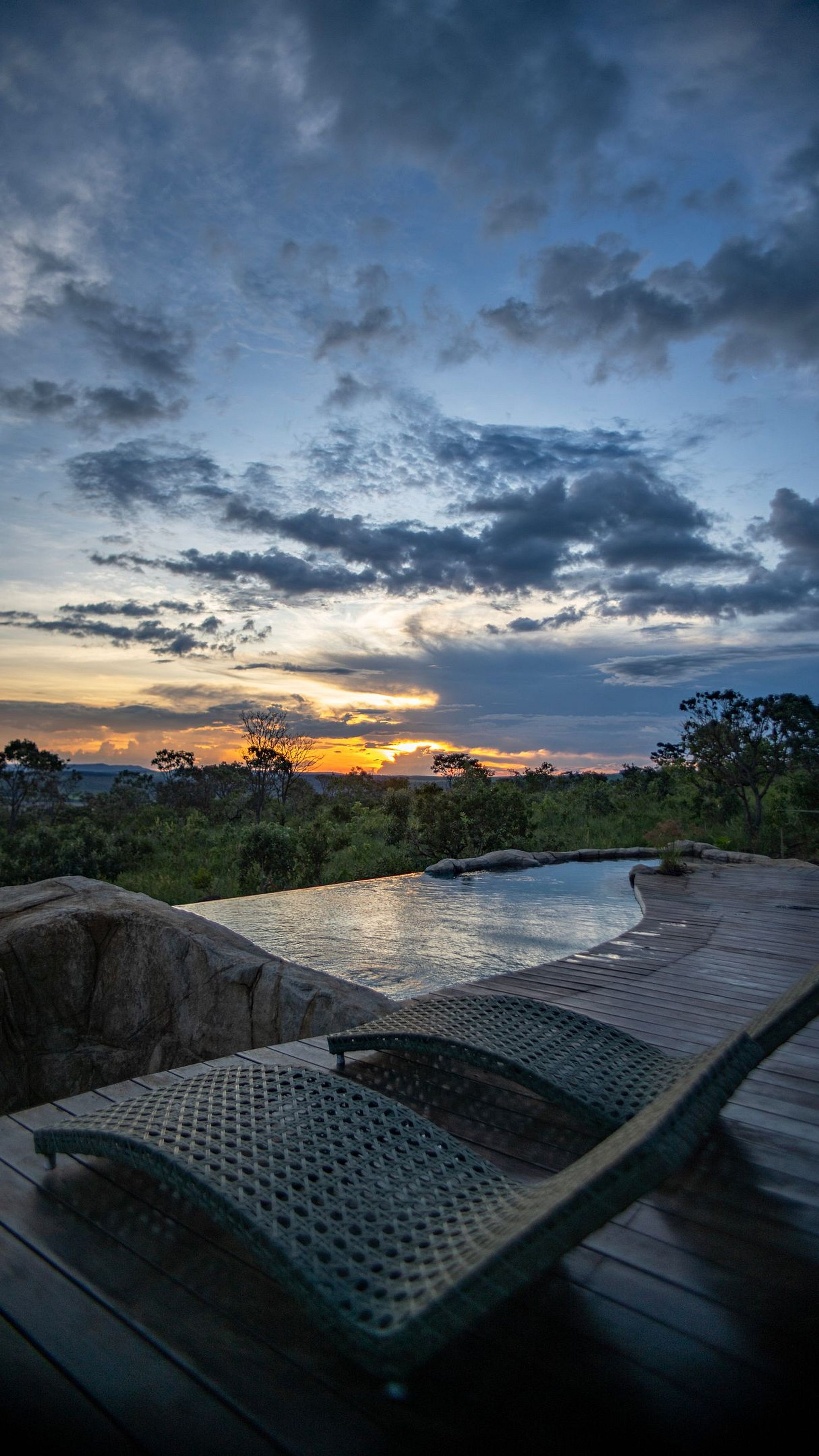 A wooden deck with lounge chairs overlooking a pool, surrounded by greenery and a sunset view.