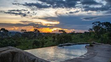 A wooden deck with lounge chairs overlooking a pool, surrounded by greenery and a sunset view.