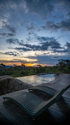 Terraza de madera con sillones de descanso con vista a una piscina, rodeada de vegetación y una vista al atardecer.