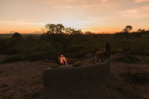 Una persona sentada junto a una fogata en un entorno natural, con árboles y un atardecer al fondo.