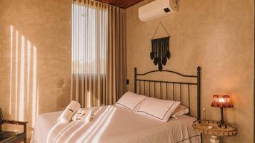 A bedroom with a bed, a bedside table with a lamp, a wall-mounted air conditioner, and a window with curtains.