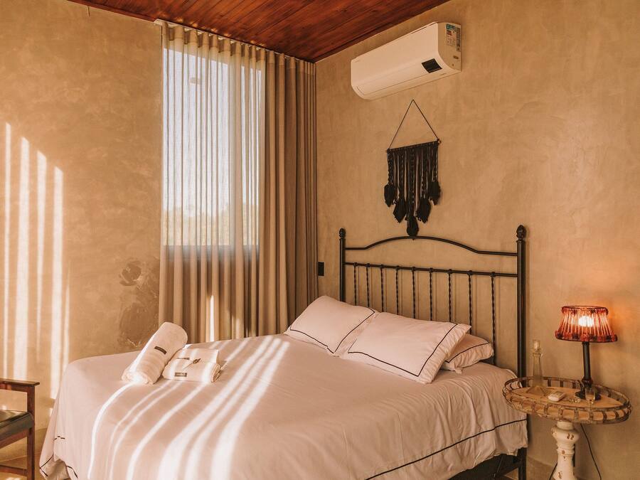 A bedroom with a bed, a bedside table with a lamp, a wall-mounted air conditioner, and a window with curtains.