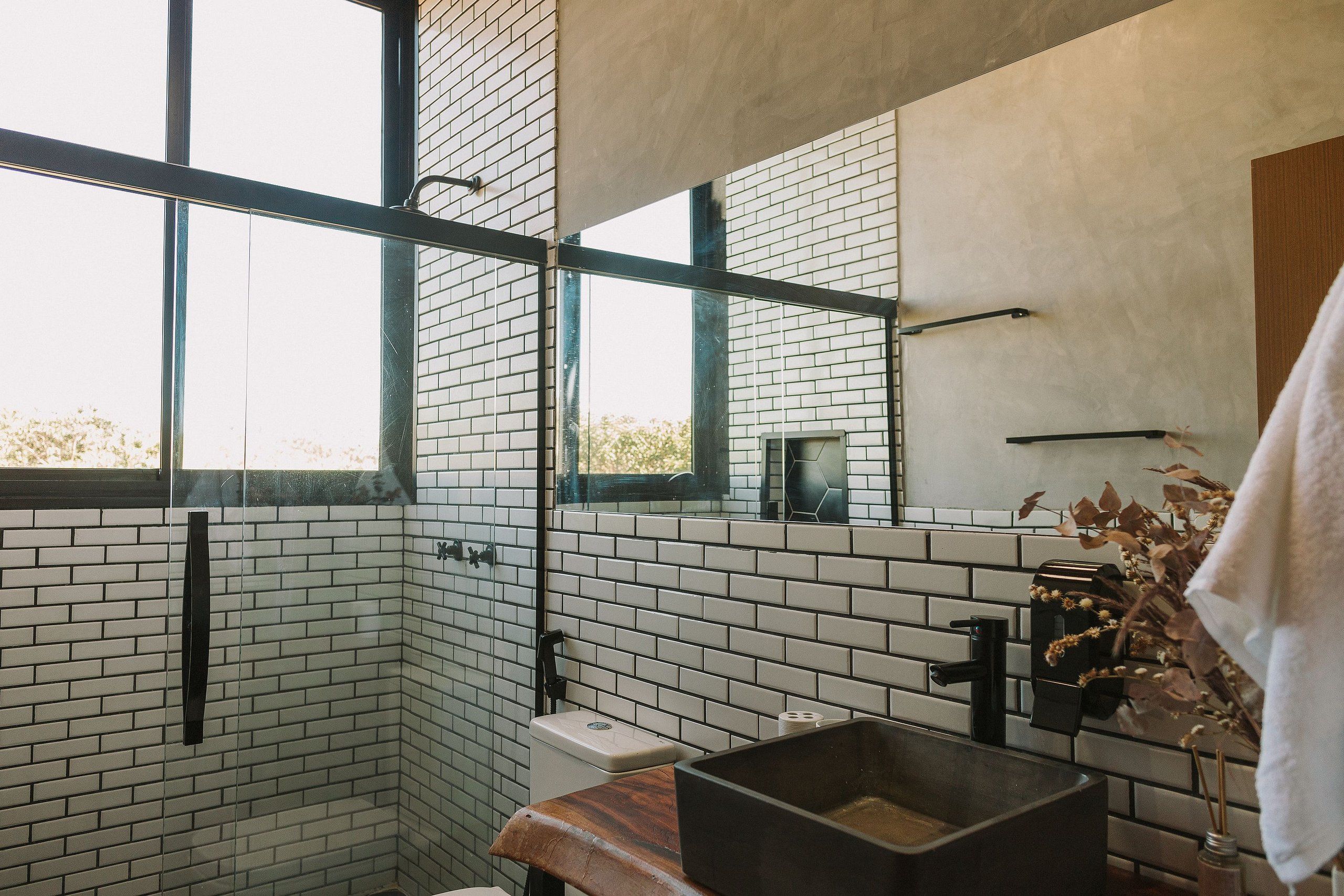 Un baño moderno con paredes de ladrillo blanco, un ventanal grande, un banco de madera y un lavabo negro.