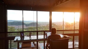 A person seated at a table with a laptop, overlooking a scenic landscape through large windows.
