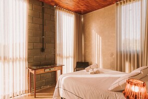 A bedroom with a bed, a nightstand, a lamp, and a brick wall. - CASA ÔNIX (Alto Paraíso de Goiás)