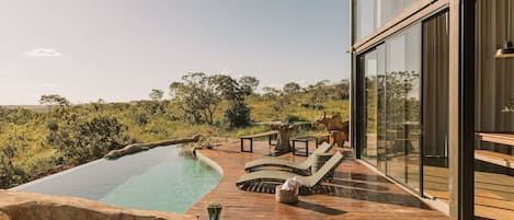 A wooden deck with a swimming pool, lounge chairs, and a glass-enclosed room.
