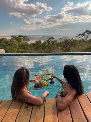 Two people at a poolside table with a spread of food and drinks, overlooking a scenic landscape. - CASA ÔNIX (Alto Paraíso de Goiás)
