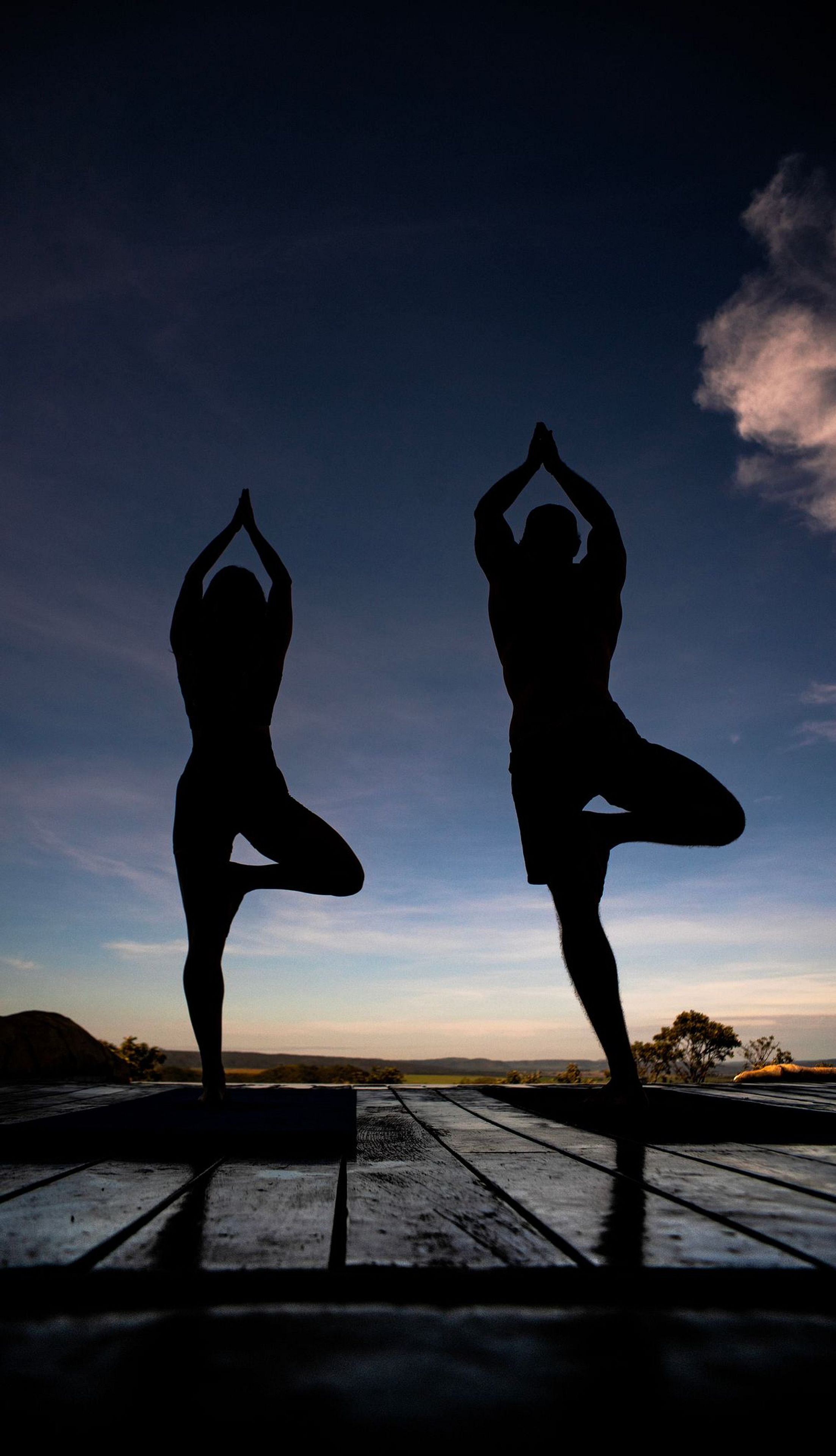 Dos siluetas de personas realizando posturas de yoga sobre una plataforma de madera al atardecer.