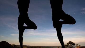 Dos siluetas de personas realizando posturas de yoga sobre una plataforma de madera al atardecer.
