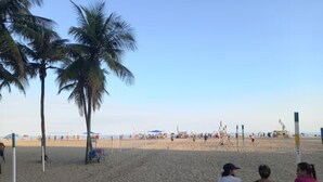 A beach with palm trees, a volleyball net, and people sitting on a bench.