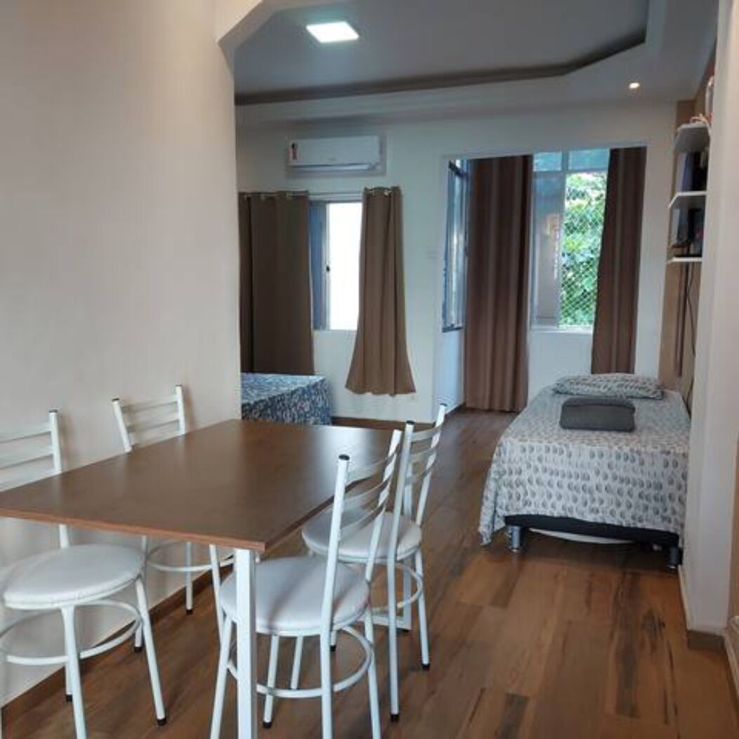A room with a wooden dining table and white chairs, a bed with a patterned bedspread, and a window with curtains.