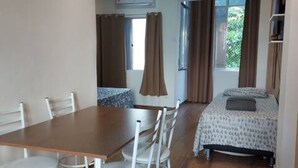 A room with a wooden dining table and white chairs, a bed with a patterned bedspread, and a window with curtains.
