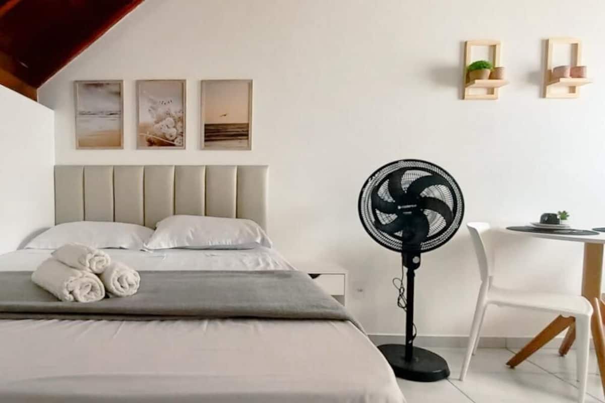 Ein Hotelzimmer mit Bett, Standventilator, kleinem Tisch mit Stuhl und Wandregalen mit Dekoration.