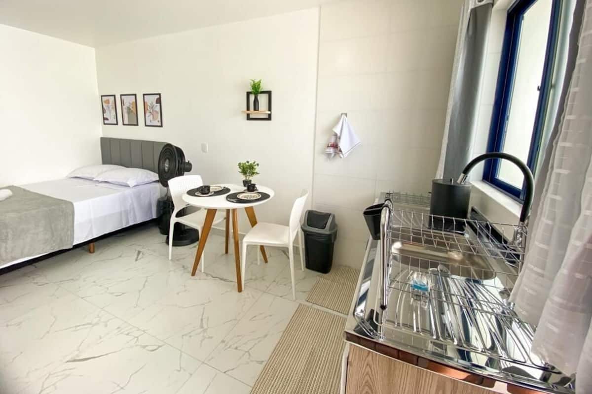 Ein kleines Hotelzimmer mit Bett, einem Esstisch mit Stühlen, einer Küchenzeile inklusive Geschirrspüler und einem Fenster mit Vorhängen.