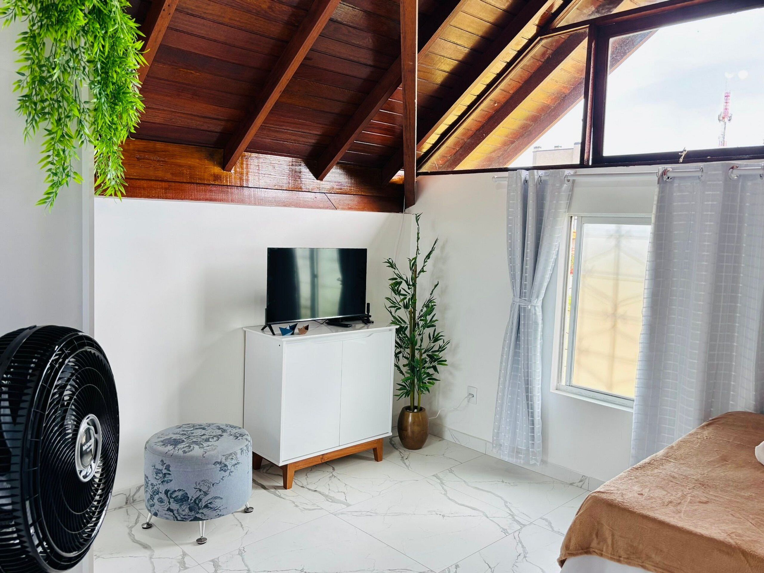 A room with a wooden ceiling, a white cabinet with a flat-screen TV, a potted plant, a large fan, a round ottoman, and a bed with a beige cover.