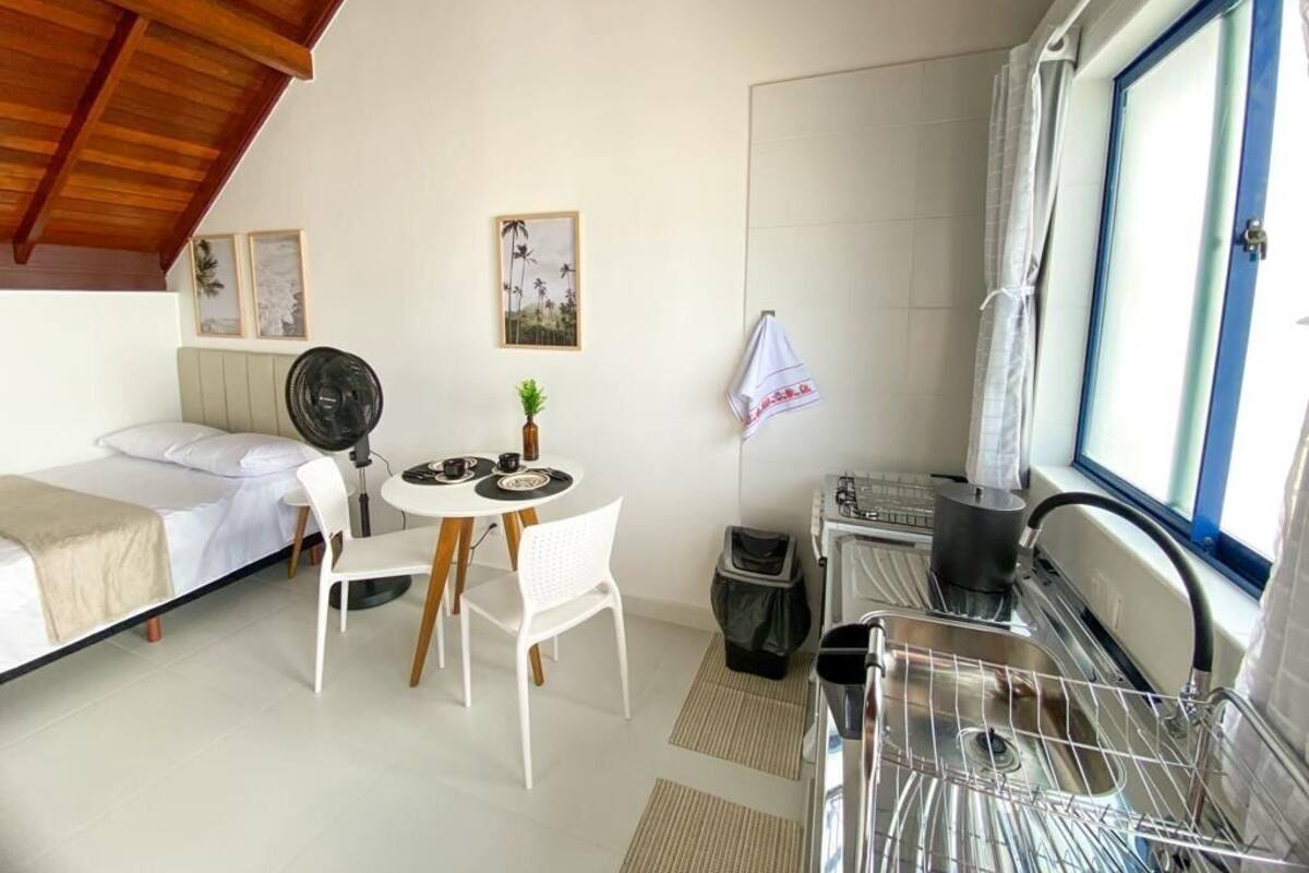 Ein kleiner Raum mit einem Bett, einem Tisch mit Stühlen, einem Ventilator, einem Fenster mit Vorhängen und einer Waschmaschine.