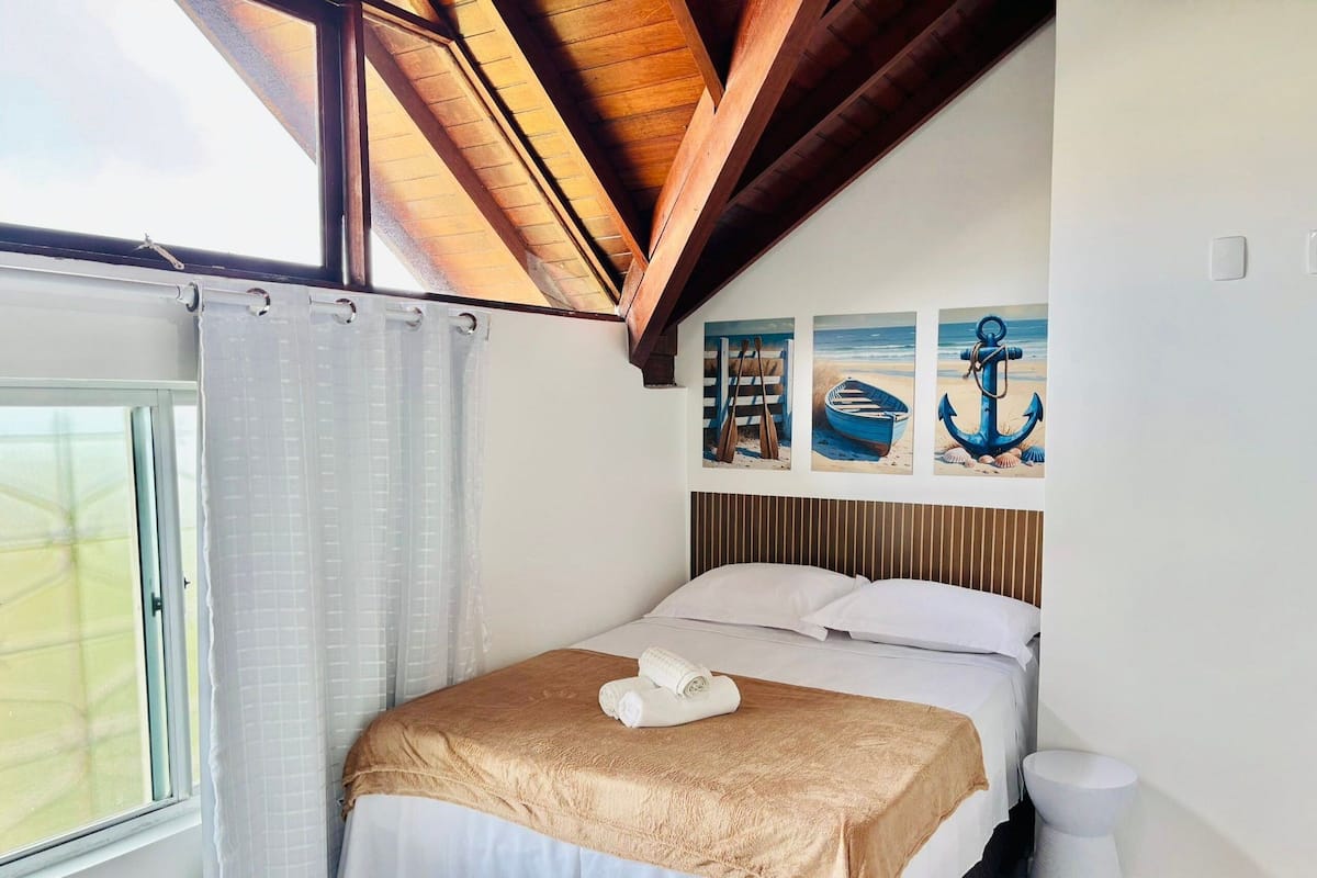 Ein Schlafzimmer mit einem Bett, einem Fenster mit Vorhängen, einer Holzbalkendecke und drei Bildern an der Wand.