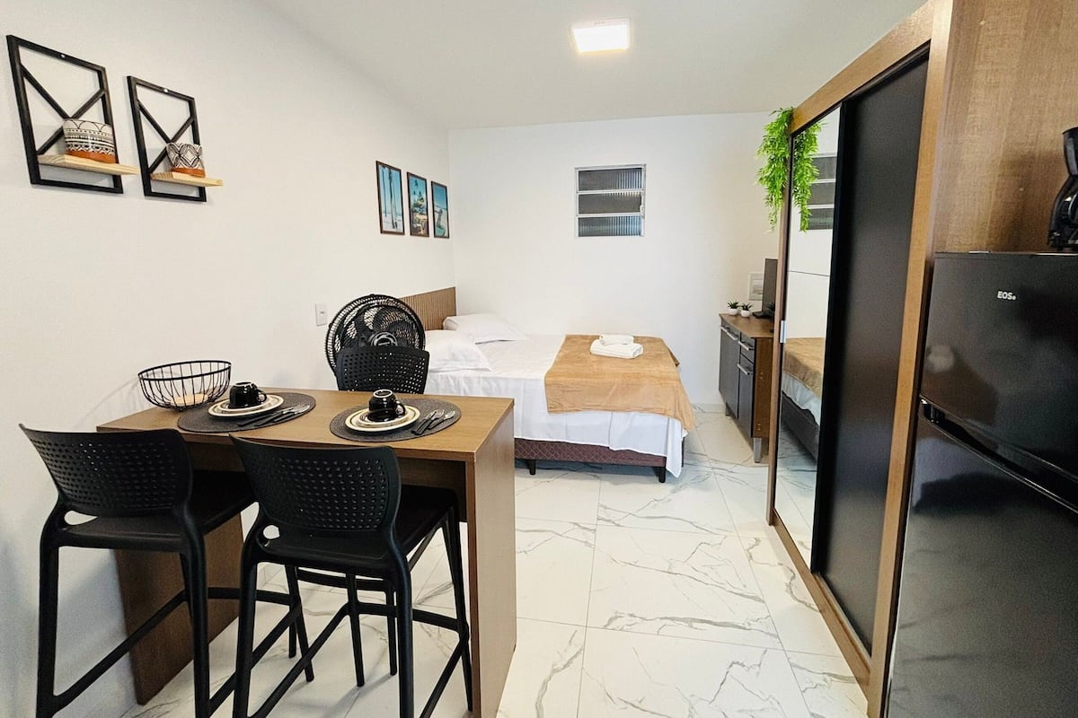 Ein kleines Hotelzimmer mit Bett, Schreibtisch, Stuhl, Esstisch und einer Küchenzeile.