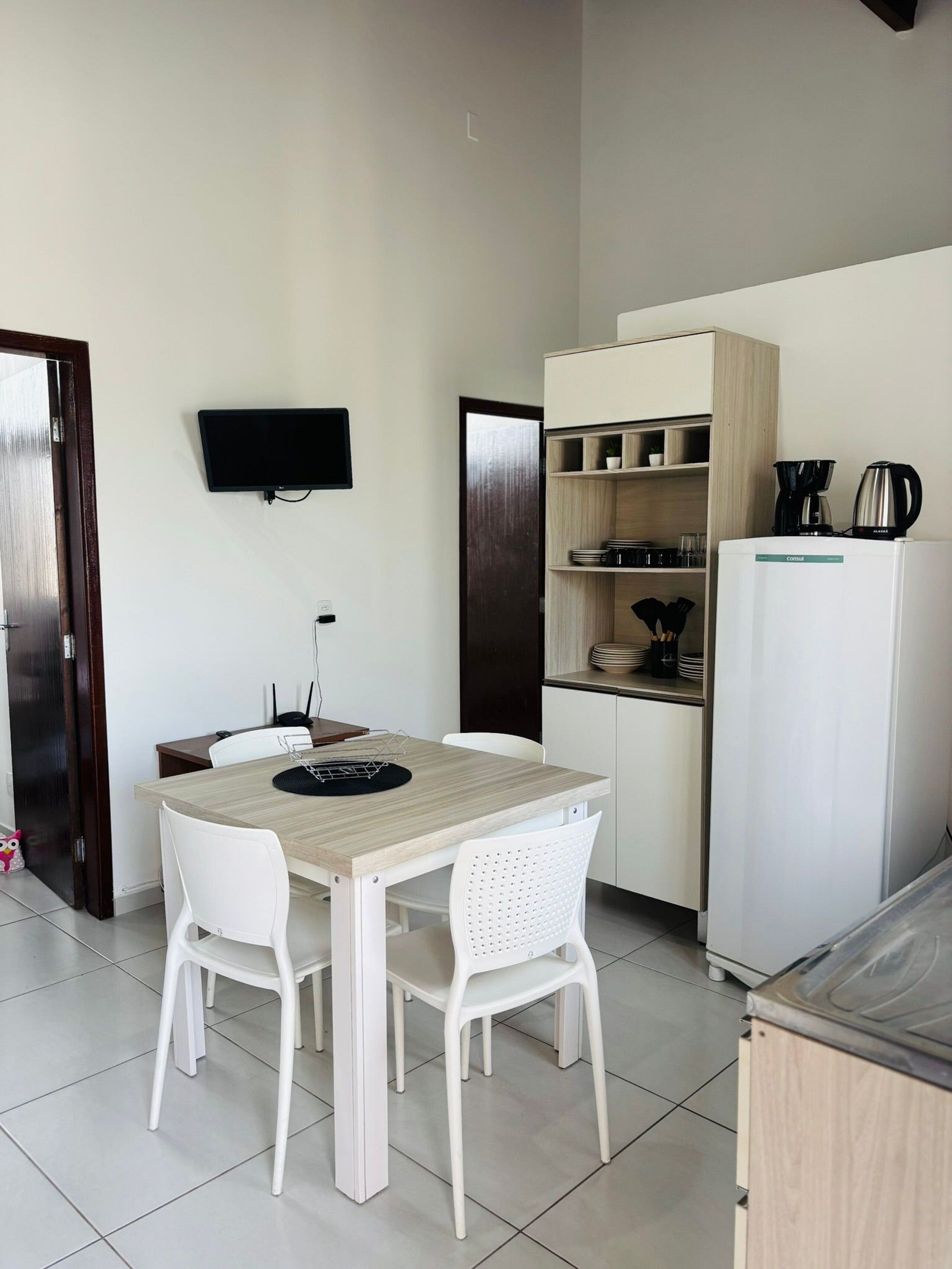 A compact kitchen with a wooden table, white chairs, a white refrigerator, and a TV mounted on the wall.