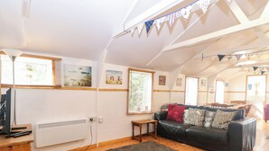 A cozy living room with a black leather sofa, a grey rug, and wooden flooring. There is a TV on a wooden stand, a small table, and a radiator. - Suzanne's Beach Hut (Norwich)