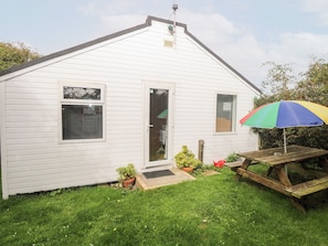Een wit huis met één verdieping, met een voordeur, twee ramen, een klein terras en een houten picknicktafel met een kleurrijke parasol.