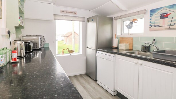 A modern kitchen with white cabinets, a black countertop, and a view of a house through a window. - Suzanne's Beach Hut (Norwich)