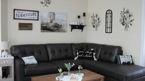 Un salon avec un canapé en cuir foncé, une table basse agrémentée d’objets décoratifs, et des éléments décoratifs muraux, notamment un panneau indiquant « Ferme » et un coussin avec l’inscription « Un jour à la fois ».
