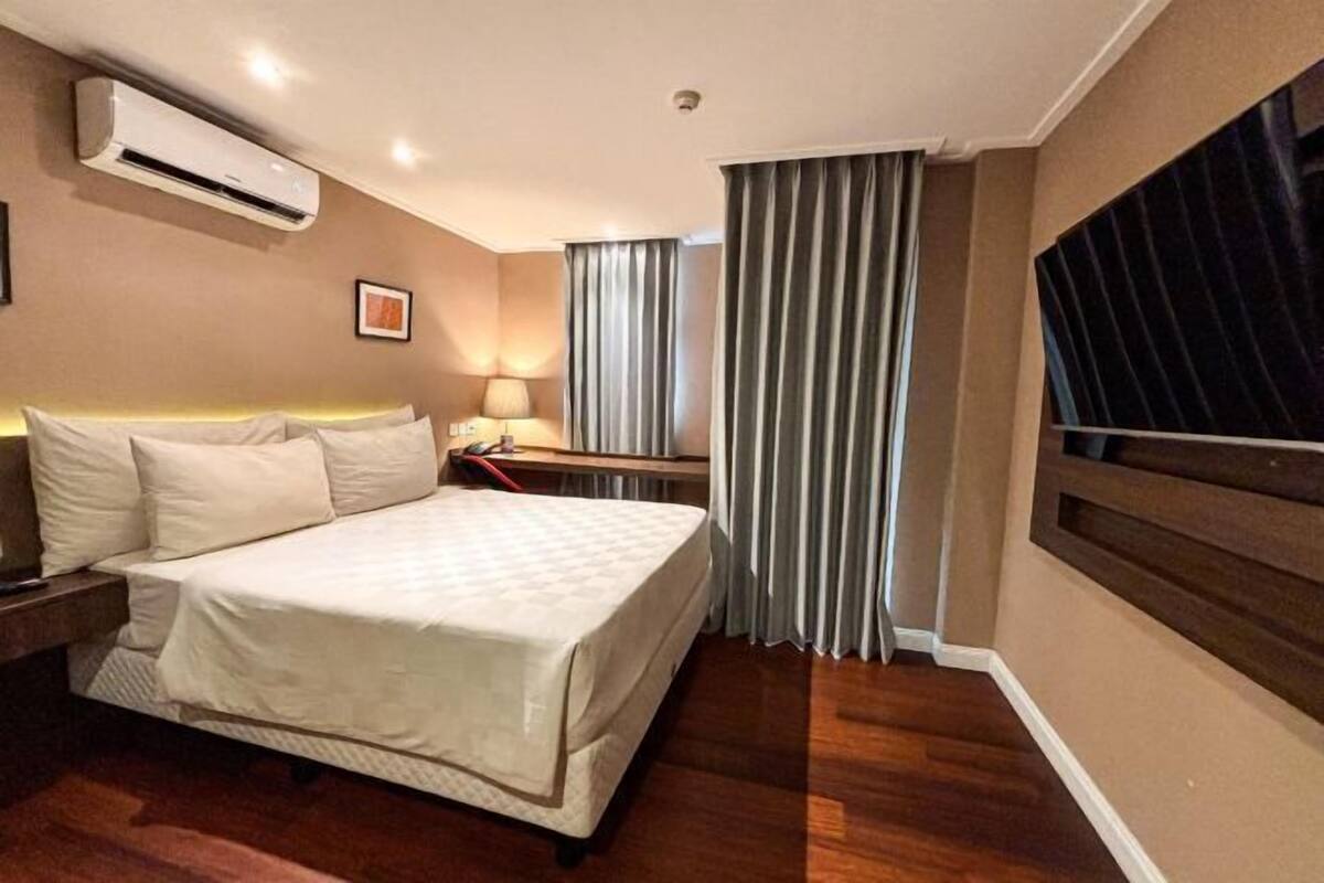 Habitación de hotel con cama, mesita de noche, lámpara, unidad de aire acondicionado, cortinas y un ventanal grande.