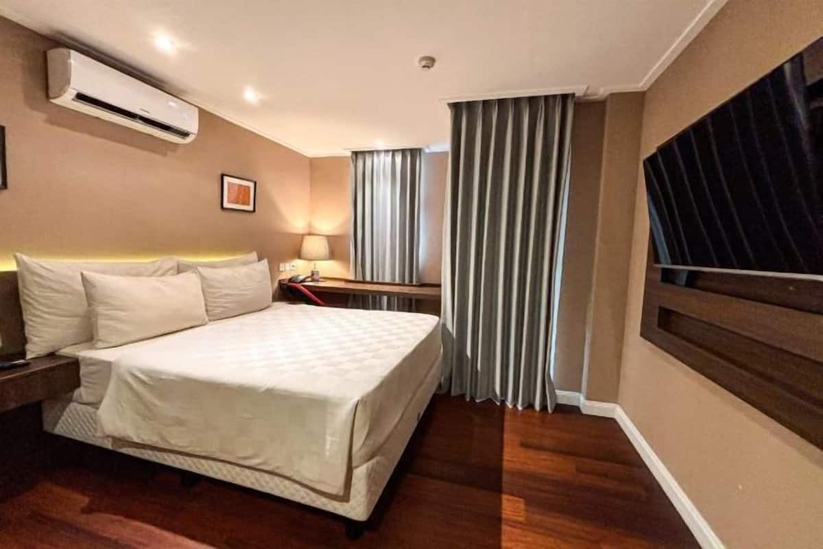 Habitación de hotel con cama, mesita de noche, lámpara, unidad de aire acondicionado, cortinas y un ventanal grande.