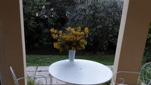 Une table ronde surmontée d’un vase blanc rempli de fleurs jaunes, entourée de chaises en métal blanc, sur une terrasse carrelée donnant sur un jardin.