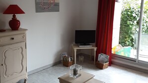 Un salon avec un téléviseur, une lampe, une table et une fenêtre avec des rideaux.